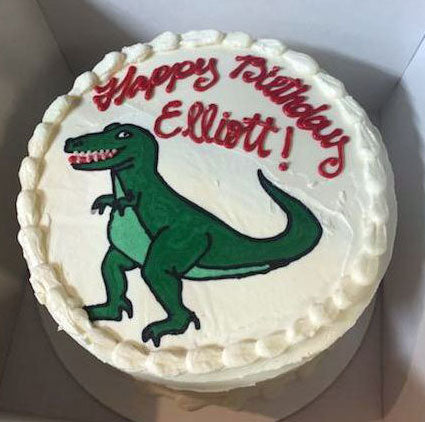A round white birthday cake with a green dinosaur in the middle and "Happy Birthday Elliott!" in red icing. 