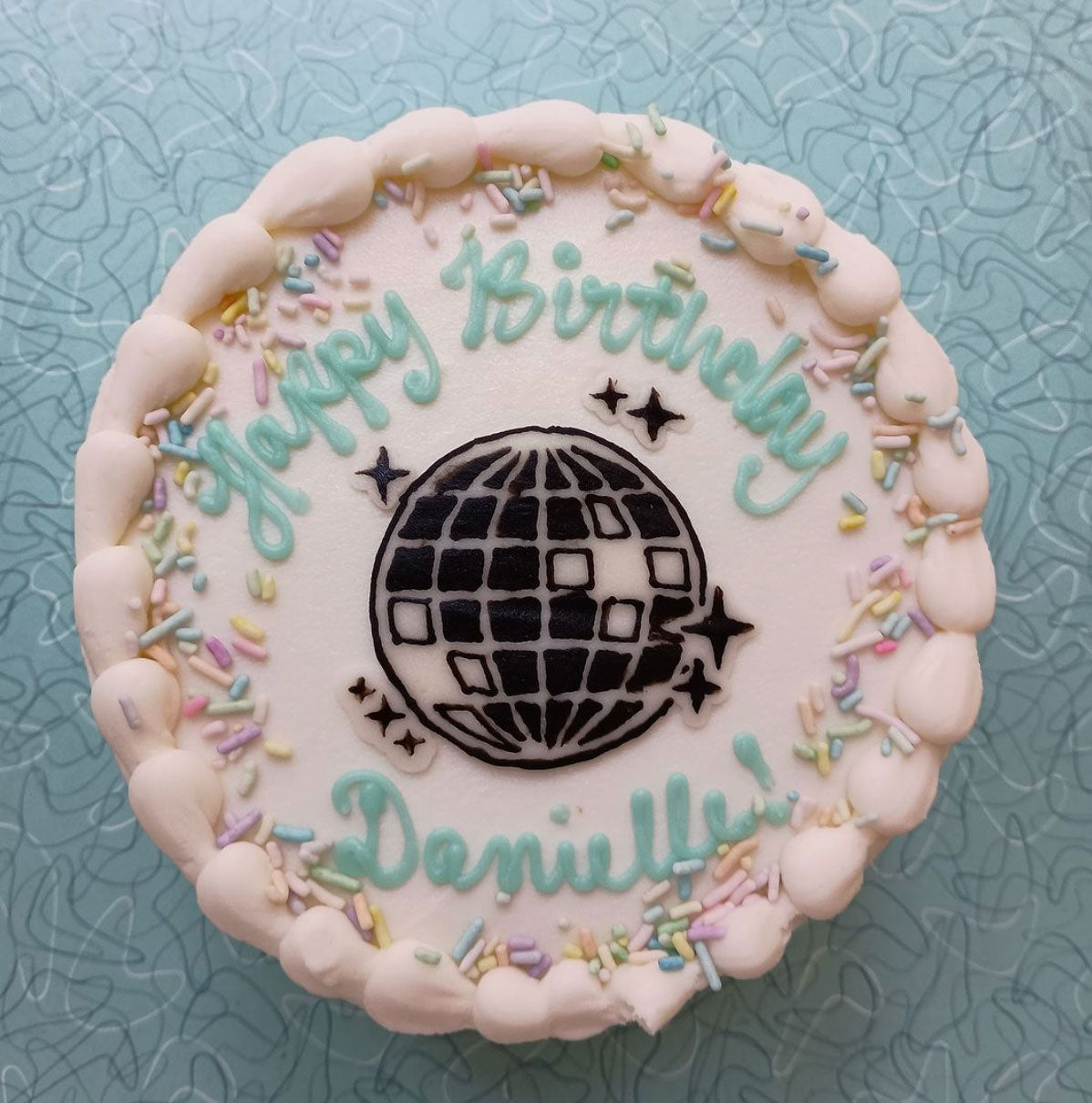 The cake is decorated with white frosting, colorful sprinkles, and a piped border. The phrase "Happy Birthday Danielle" is written in light blue icing on the top. A disco ball design is prominently featured in the center of the cake, surrounded by star accents.