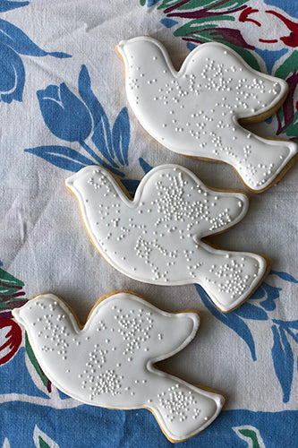Three dove-shaped sugar cookies are decorated with smooth white royal icing and topped with small white nonpareil sprinkles. The cookies are arranged diagonally on a floral-patterned fabric featuring blue, red, and green tulip motifs. The dove cookies symbolize peace and are ideal for events such as weddings, baptisms, or holidays. The overall presentation has a soft, homemade charm perfect for festive or meaningful occasions.