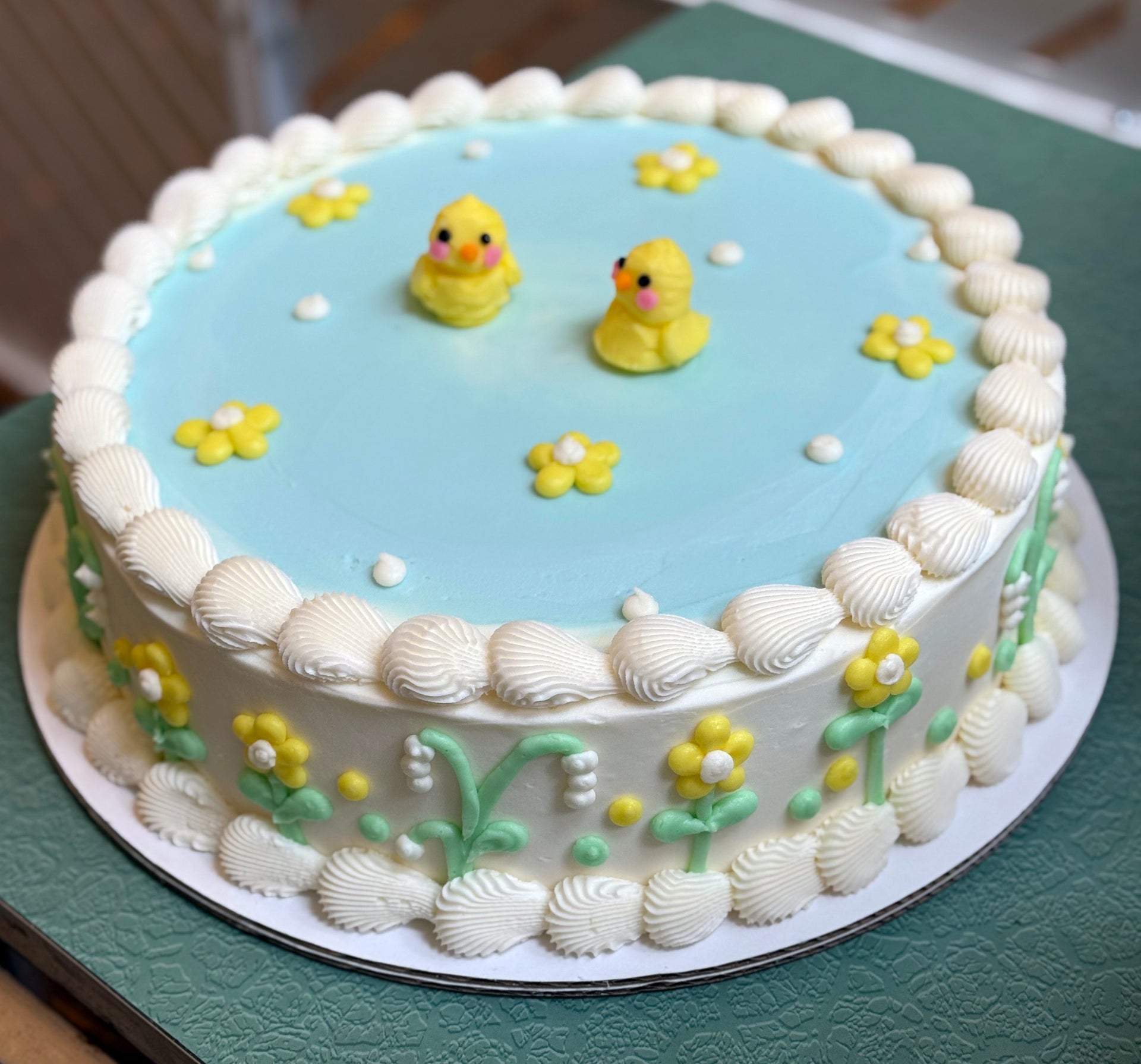 A decorated cake with white buttercream frosting and blue buttercream, adorned with yellow daisy-like flowers and small sugar ducks.