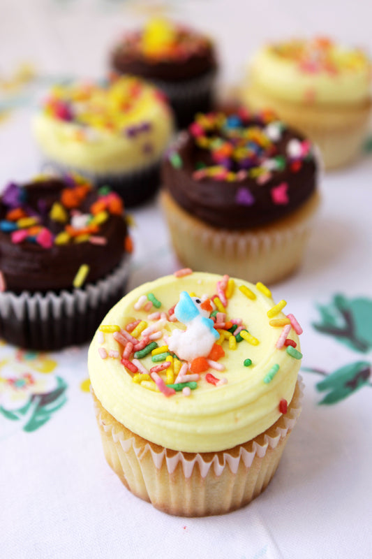 A selection of cupcakes with one frosted in white buttercream and topped with multicolored sprinkles, and another in dark chocolate frosting with a single white chocolate pearl on top. Both are on a paper liner with floral patterns.
