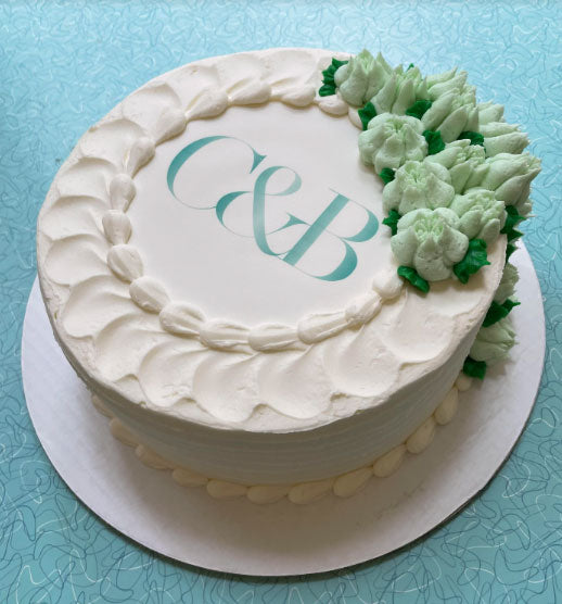 White wedding or celebration cake with "C&B" initials in teal on top, adorned with light green buttercream floral decorations on a blue table.