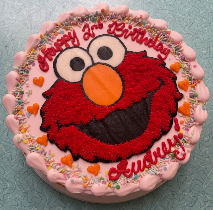 Round birthday cake with red Elmo character and colorful sprinkles around the border, decorated with 'Happy 2nd Birthday Audrey' in red icing.