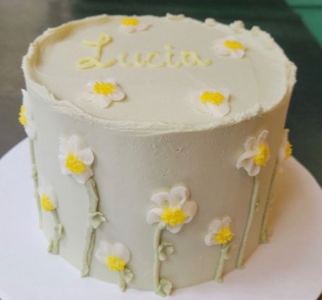 A light green frosted round cake decorated with white and yellow daisy flowers and the name "Lucia" written in yellow script on top.