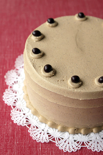 A round chocolate cake with espresso buttercream frosting and chocolate-covered espresso beans garnishing the top, presented on a white doily.