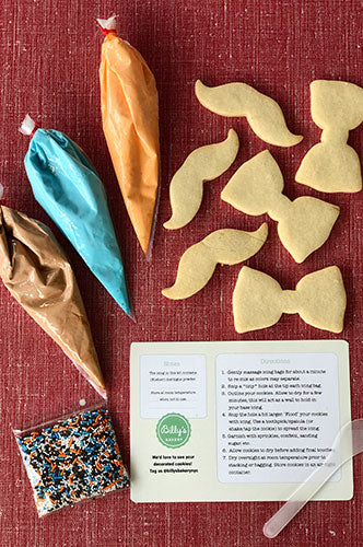 Cookie decorating kit with mustache and bow tie sugar cookies, orange, blue, and brown icing bags, colorful sprinkles, and decorating instructions on a red background.