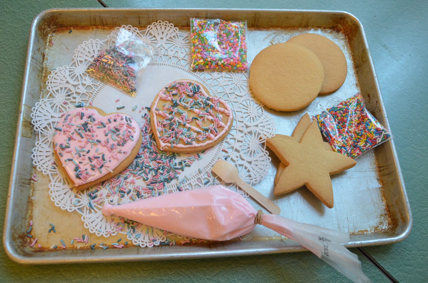 Heart-shaped sugar cookies decorated with pink icing and colorful sprinkles on a baking sheet, alongside a piping bag of pink frosting.