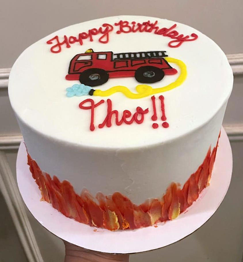 Round white birthday cake with a red Fire truck in the middle and "Happy Birthday Theo!!". Red flames are around the bottom.