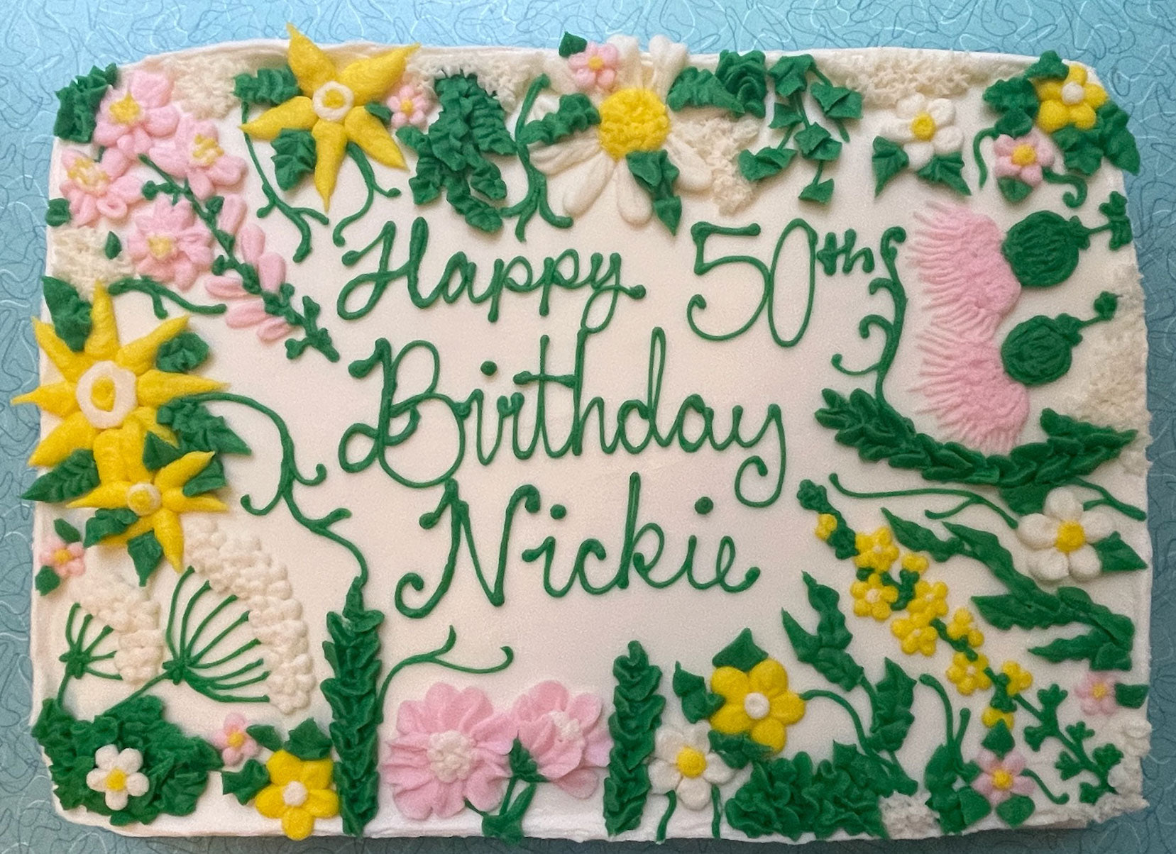 White birthday cake with green 'Happy 50th Birthday Nickie' text, adorned with yellow, pink, and white floral decorations and green leaves.