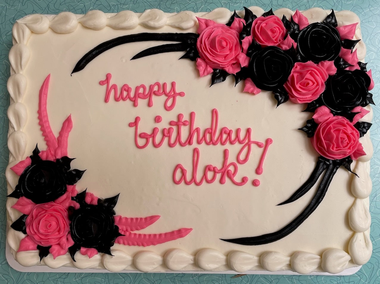 A rectangular white birthday cake decorated with pink and black frosting. The cake features pink and black roses and ribbons in the top right and bottom left corners. The center of the cake has "happy birthday alok!" written in pink script frosting. The edges of the cake are piped with white frosting and the cake sits on a blue table.