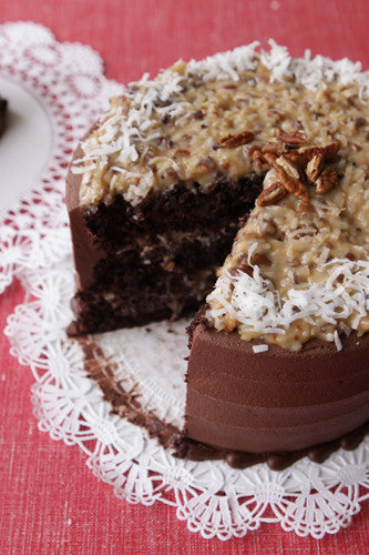 A chocolate cake with coconut pecan topping and chocolate frosting, on a cake board, with a slice taken out, placed on a red surface with a lace doily.