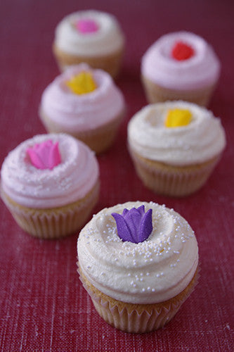 Six frosted cupcakes, mostly vanilla with white or light pink frosting, each topped with a colorful flower candy decoration and white nonpariel garnish.