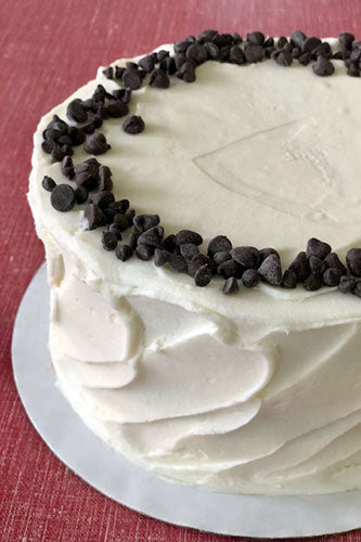 A round chocolate chip cake with white vegan buttercream frosting garnished with chocolate chips around the edge, placed on a cake board. 