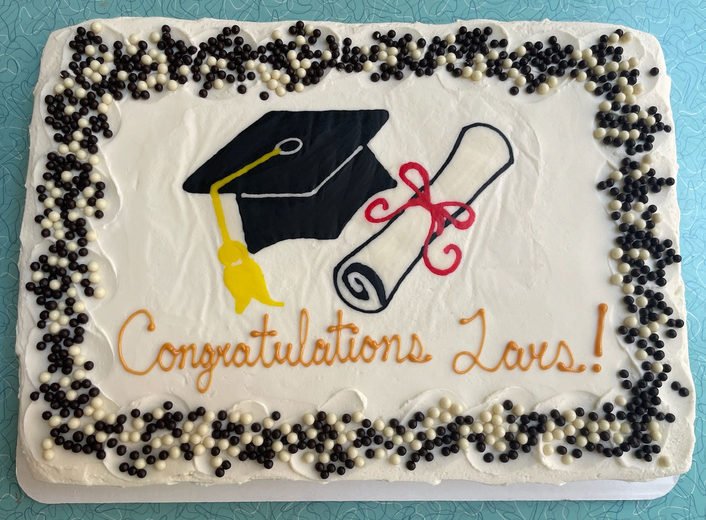Rectangular white graduation cake decorated with a black graduation cap, a yellow tassel, a rolled diploma with a red ribbon, and the words 'Congratulations Lars!' in orange icing, bordered by black and white sprinkles."