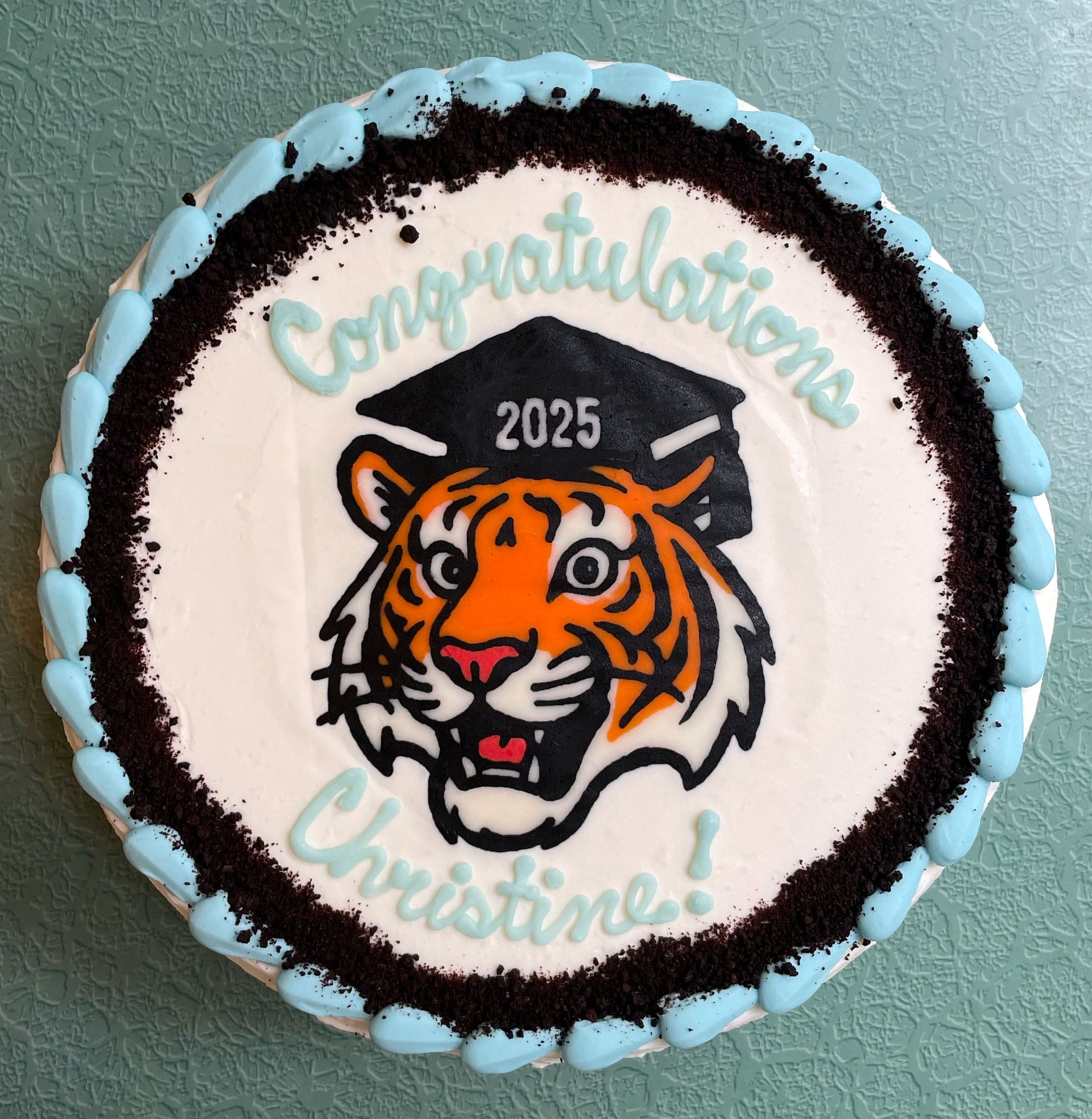 Cake with a tiger design, graduation cap, and 'Congratulations 2025 Christine!' text on a light blue background.
