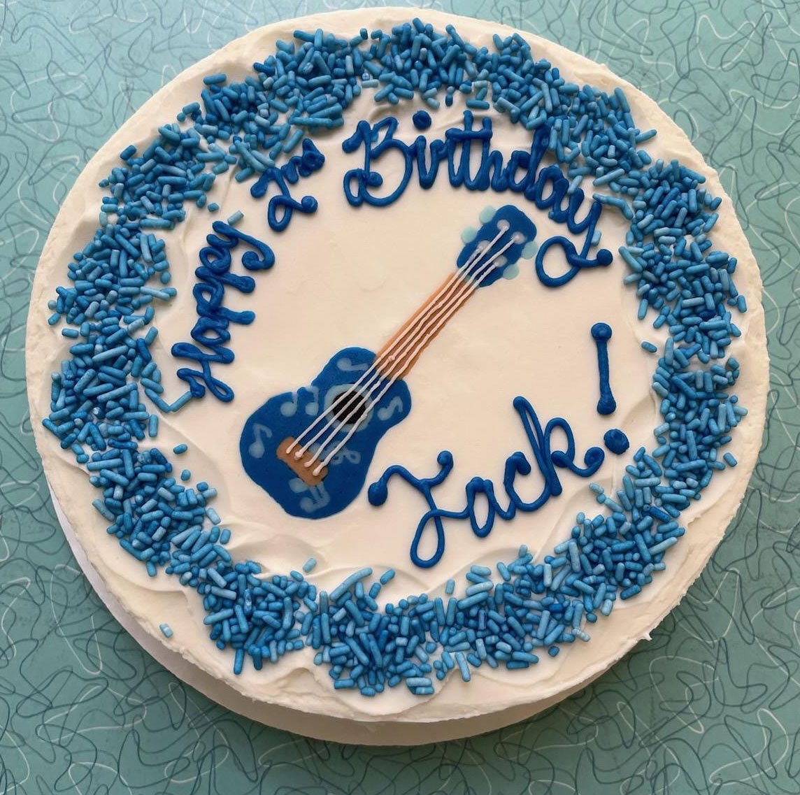 A round white birthday cake on a blue table decorated with blue sprinkles and a blue guitar in the center. The cake reads "Happy Birthday Jack!" in blue icing.