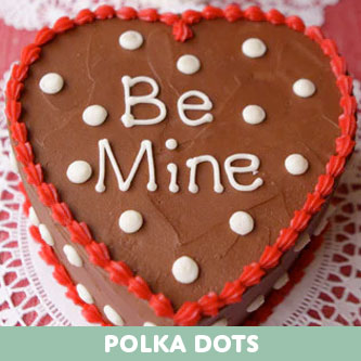 A heart-shaped chocolate cake with red piped borders and white polka dots, featuring "Be Mine" in white icing, on a white lace doily.