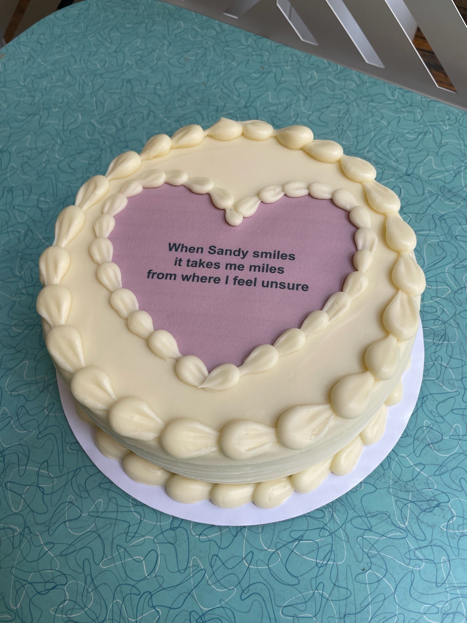 A heart-shaped cake with white frosting and a light purple heart-shaped center on a blue table. The center has text that reads: "When Sandy smiles it takes me miles from where I feel unsure."