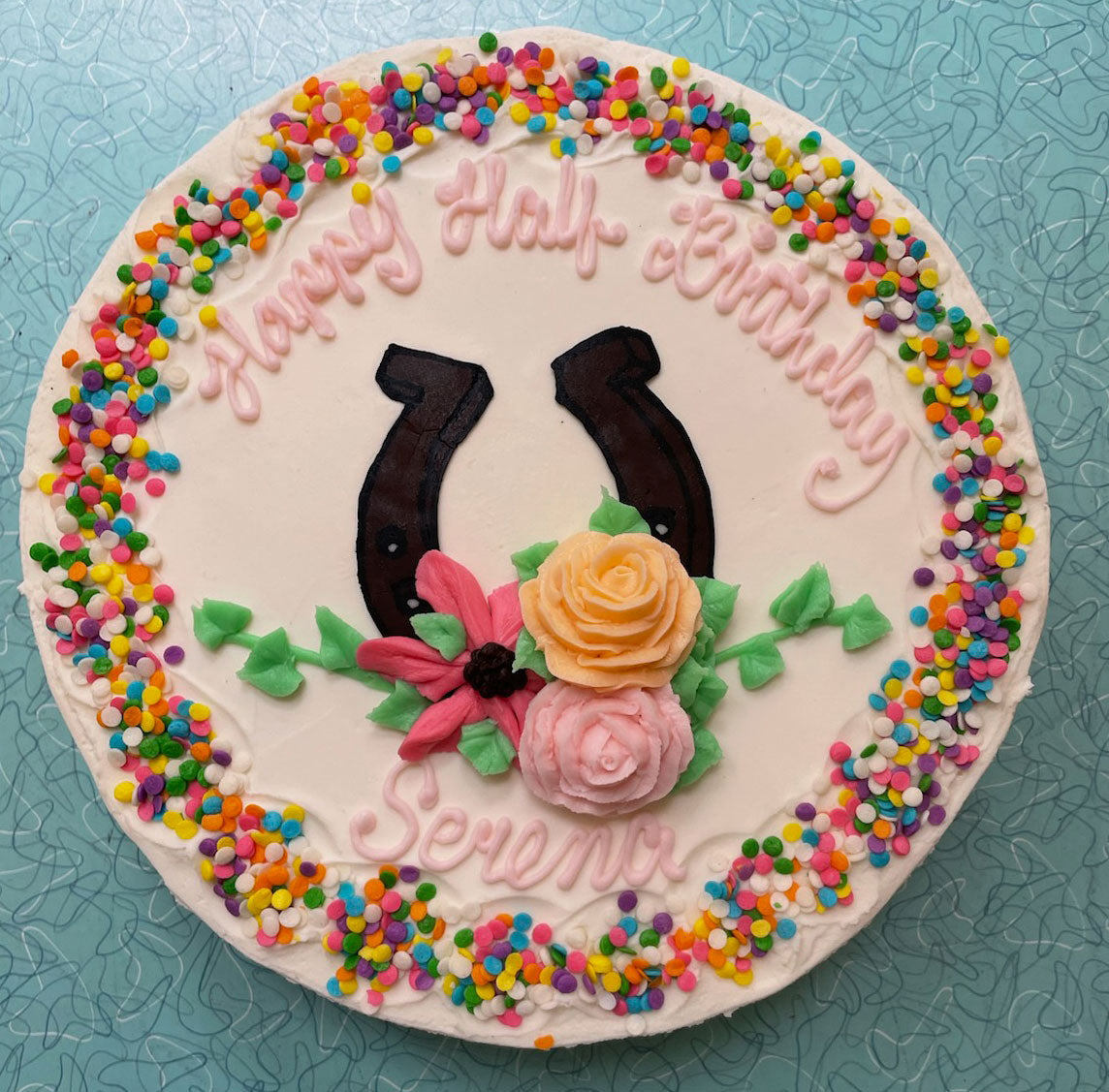 A white round cake decorated with colorful sprinkles, pink frosting text reading "Happy Half Birthday Serena," two black horseshoes, and pink and orange buttercream flowers.