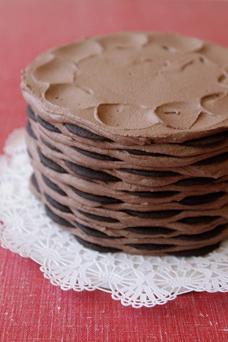 Layers of dark chocolate wafer cookies and creamy chocolate frosting stacked to form a round chocolate icebox cake on a white doily.