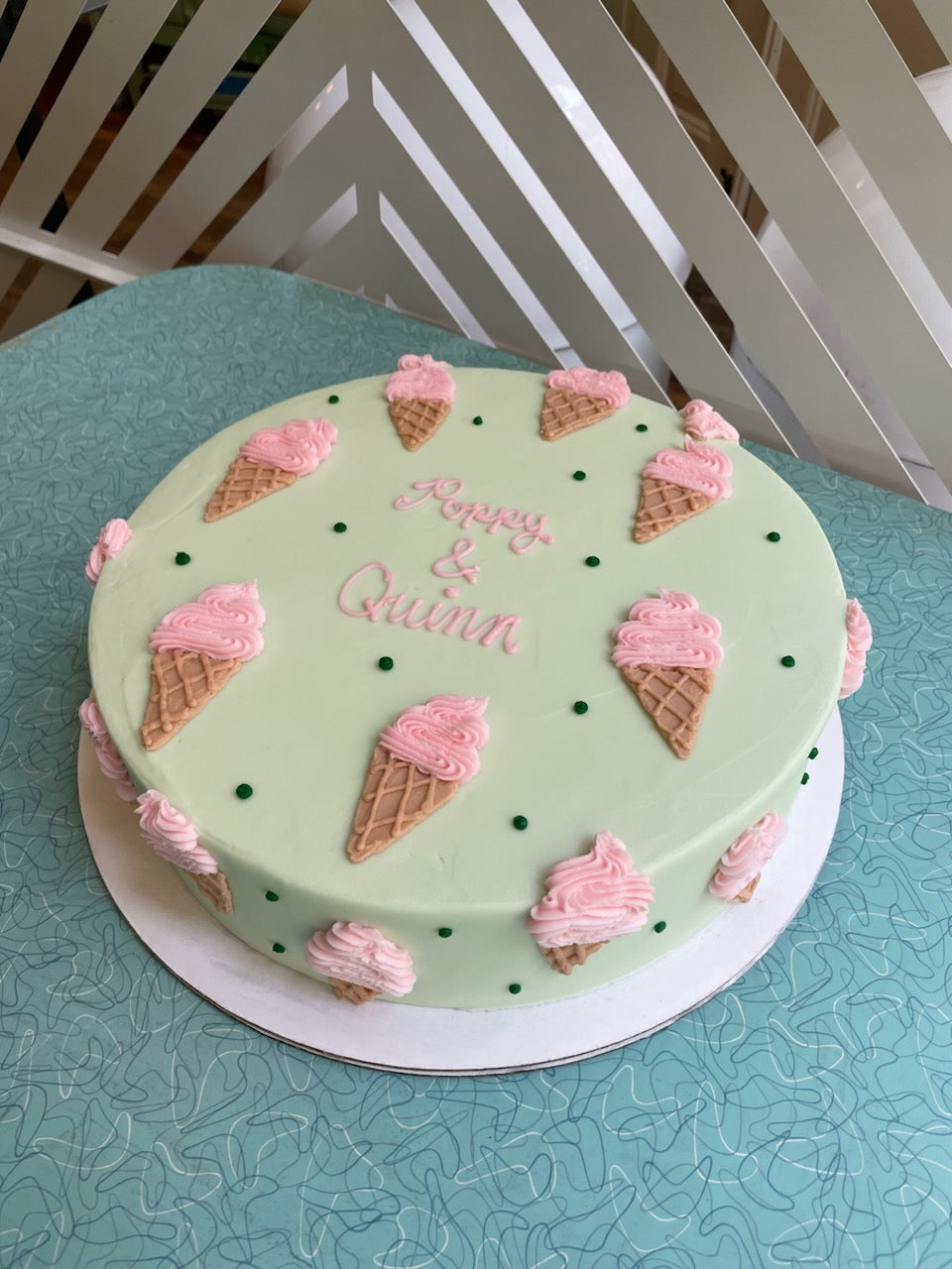 A light green round cake decorated with pink ice cream cones, green sprinkles, and the text "Happy & Quinn" in pink frosting on a blue table.