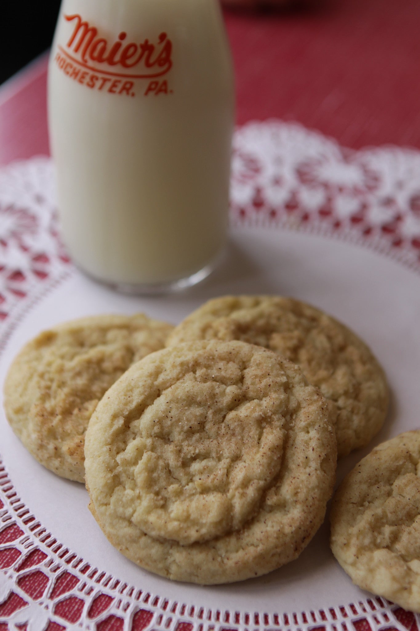 A plate of snickerdoodle cookies with a bottle of milk in the background, placed on a red and white checkered tablecloth.