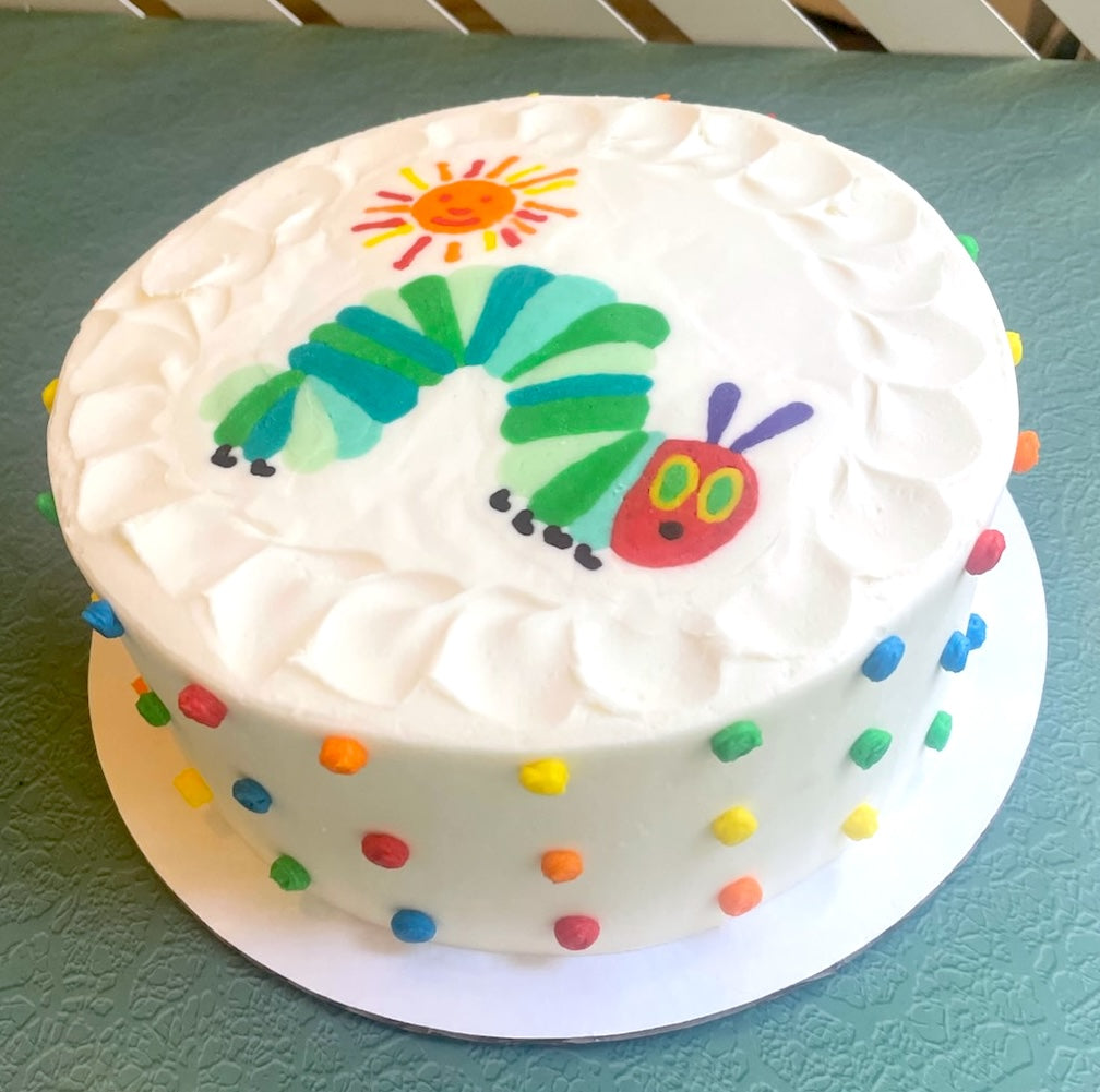 This charming image features a round, white frosted cake, decorated with multi colored dots. The top of the cake showcases a hand-drawn image depiction of "The Very Hungry Caterpillar" from Eric Carle's iconic book. The caterpillar is rendered in vibrant shades of green and blue for its segmented body, with a bright red head, purple antennae, and yellow eyes. Above the caterpillar, a cheerful orange and yellow sun shines down. The white frosting on top has textured swirls. 