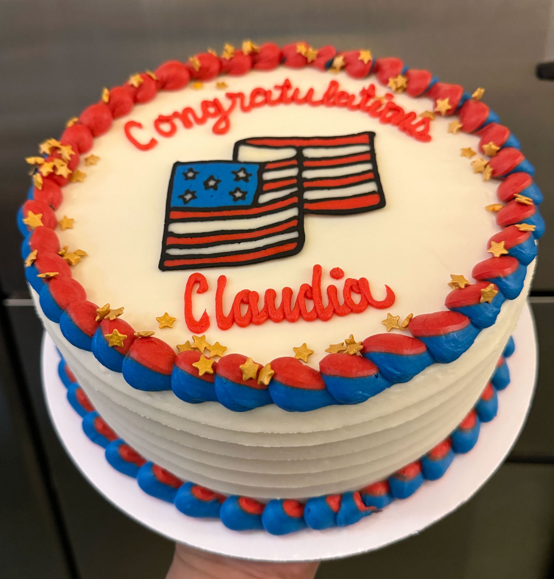 A white celebration cake decorated with red and blue frosting, featuring two American flags and the text 'Congratulations Claudia' in red script.
