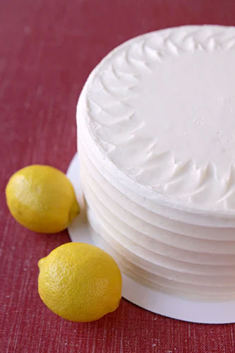 A frosted lemon cake with cream cheese icing, alongside two lemons placed on a red surface.