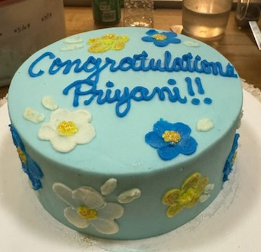 Light blue cake decorated with white, yellow, and blue flowers, with "Congratulations Priyani!!" written in blue icing on top.