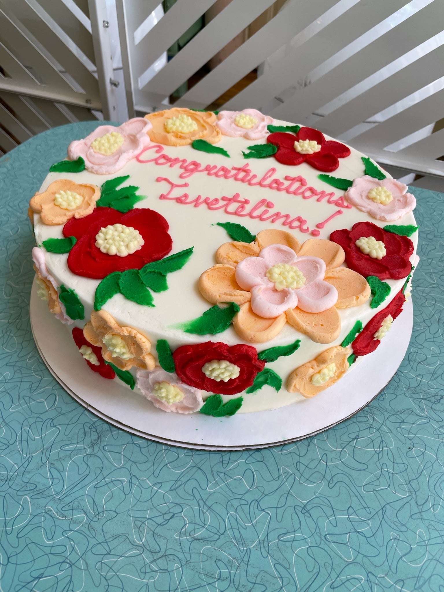 A white round birthday cake decorated with colorful buttercream flowers in shades of red, pink, orange, and light yellow, with green leaves. The top of the cake has pink frosting text that reads "Congratulations Levetelina!".