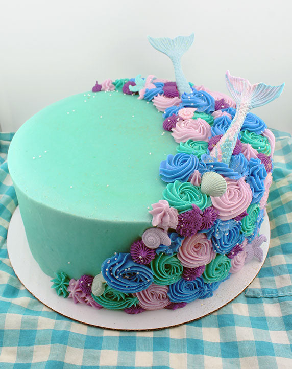A decorated cake with blue icing and purple and blue rosettes, with fondant mermaid tail and starfish decorations on top, served on a blue and white checkered tablecloth.
