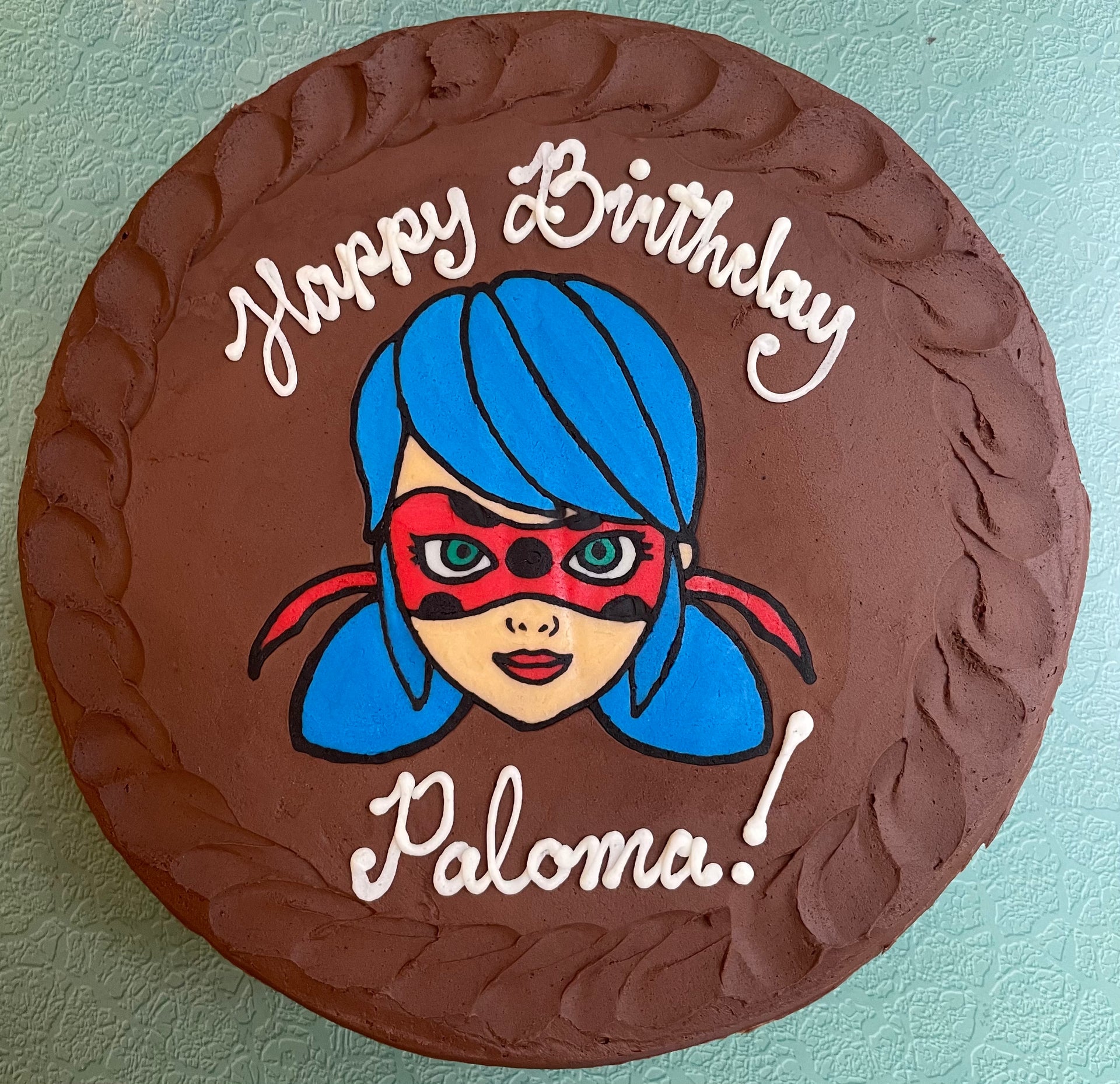 Photo of a cake frosted in chocolate buttercream with a hand-drawn image of Miraculous LadyBug. 