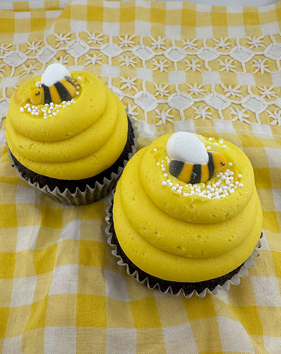 Two cupcakes with yellow frosting, decorated with edible bees and white nonpareils, on a yellow checkered background.