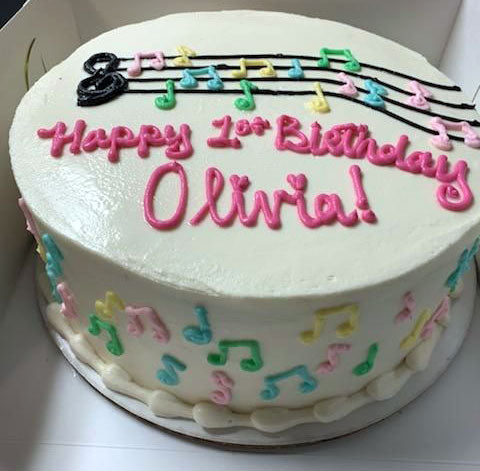 White frosted round cake decorated with colorful musical notes and a black treble clef, with "Happy 1st Birthday Olivia!" written in pink icing.