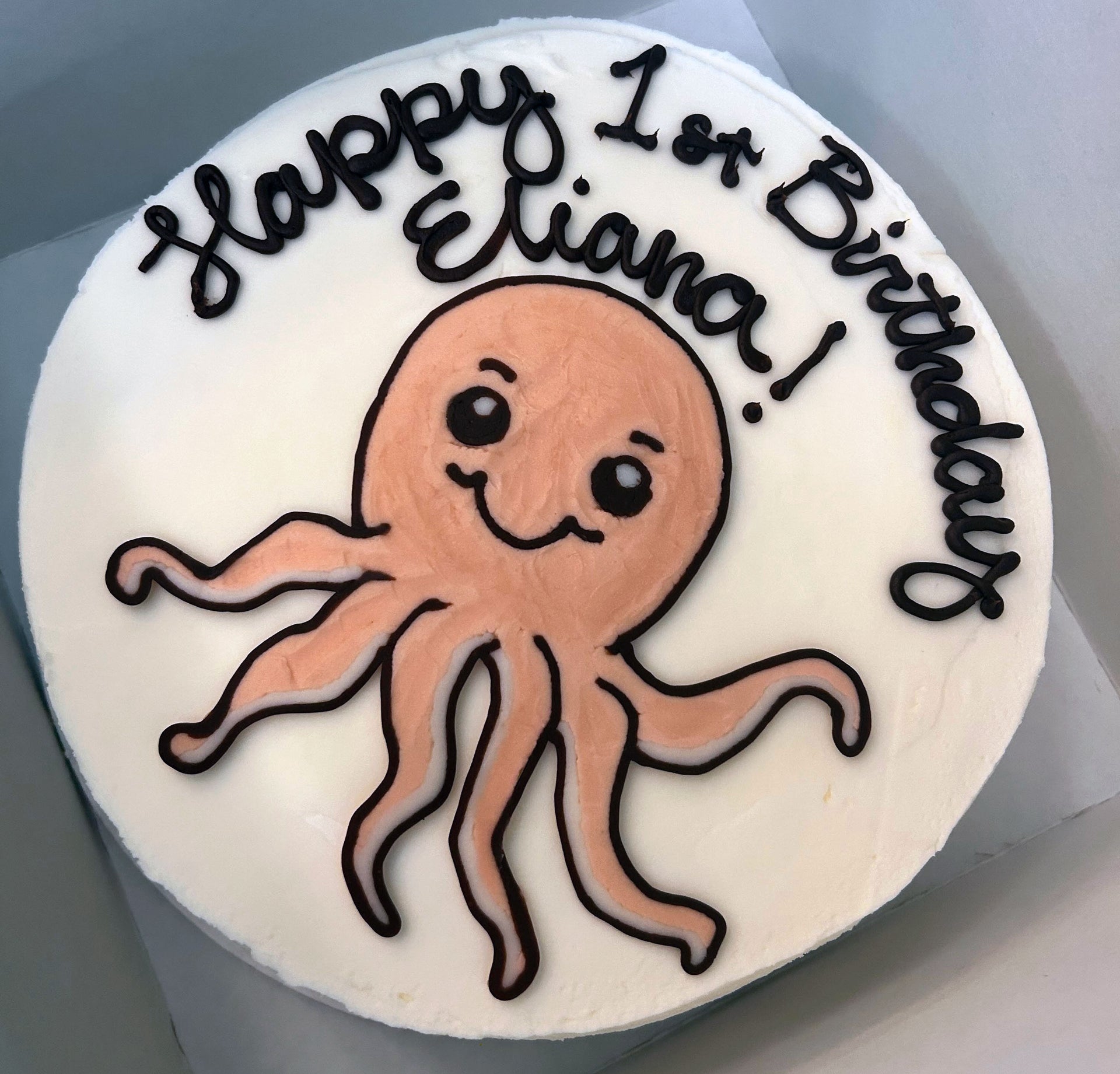 White round birthday cake with a cartoon pink octopus and black icing text that reads 'Happy 1st Birthday Eliana!'