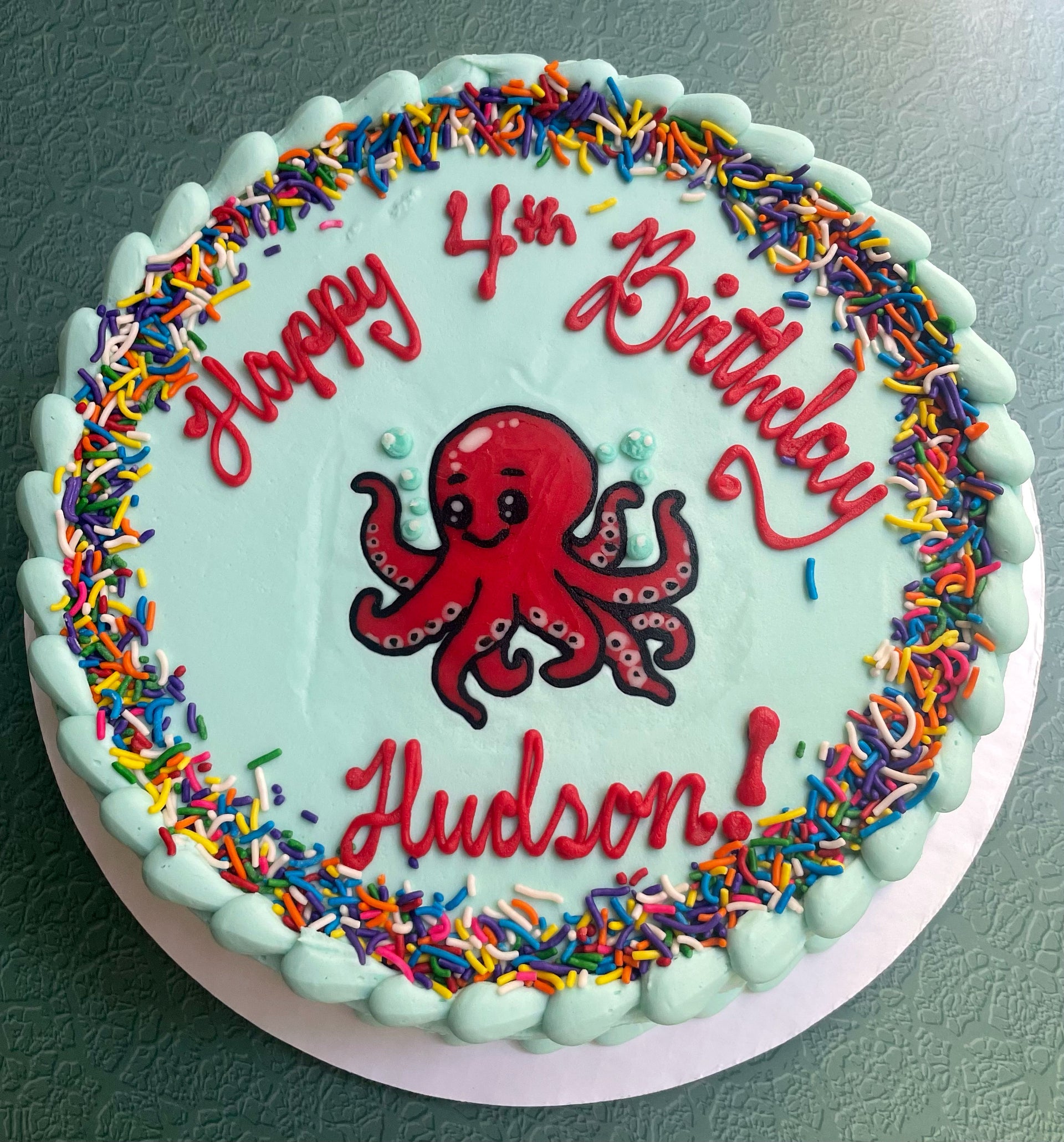 A round pastel blue cake with a red Octopus on top and rainbow sprinkles around the edges.   Birthday message on the top.