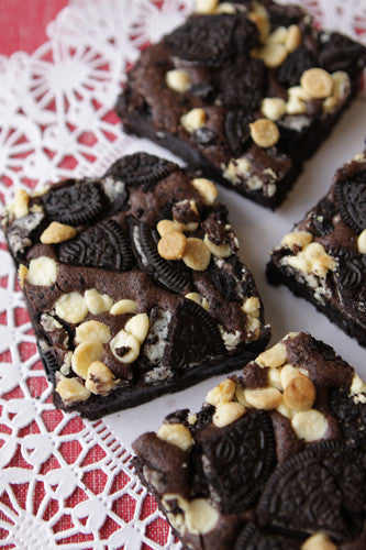 Sliced chocolate brownies topped with Oreo cookie pieces, white chocolate chips, and milk chocolate chips, presented on a plate with a lace-like pattern.