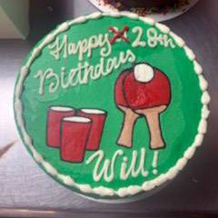 Green round birthday cake with "Happy 28th Birthday Will!" and decorations of two red paddles with a white ball, and three red solo cups, representing a beer pong or table tennis theme.