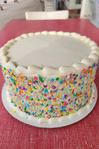 A round cake with white buttercream frosting and decorated with multicolored sprinkles on the sides on a red table.