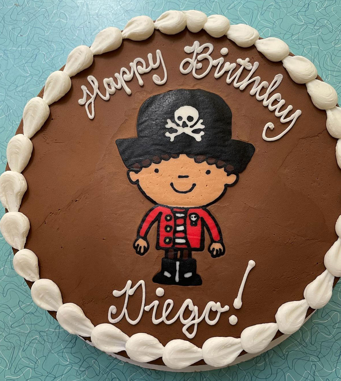A round chocolate birthday cake with white frosting border. The cake features a cartoon illustration of a pirate character. White icing text on the cake reads "Happy Birthday" above the pirate and "Diego!" below the pirate.