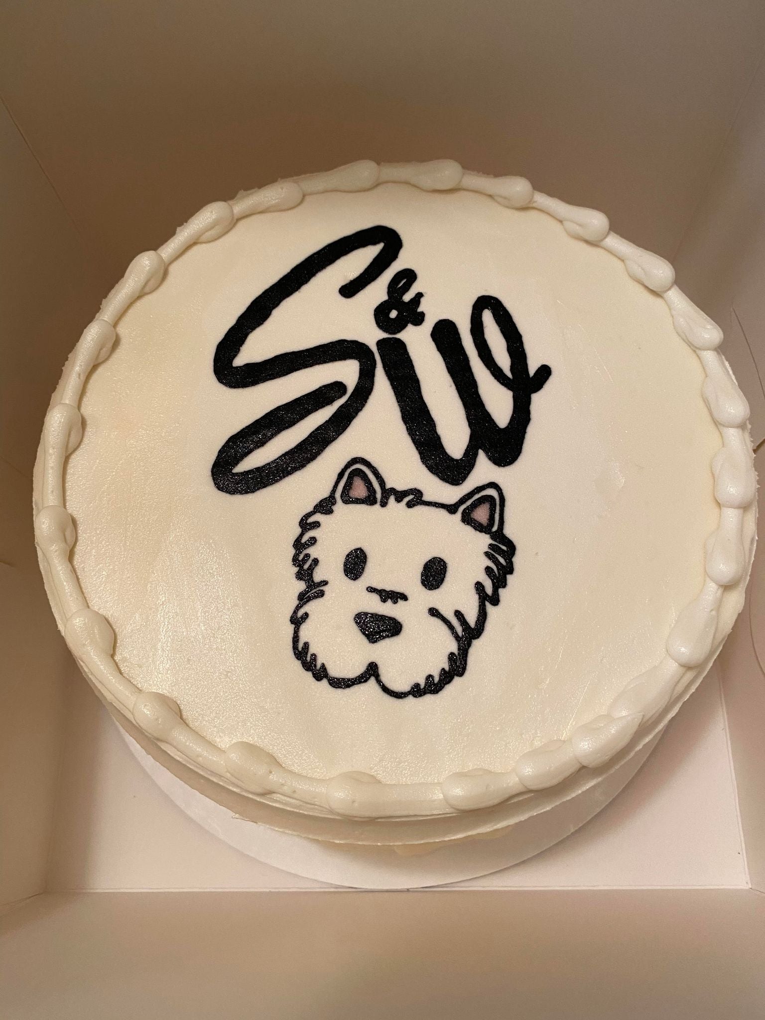 Round cake with white frosting, black text, and a small dog illustration on a plain background.