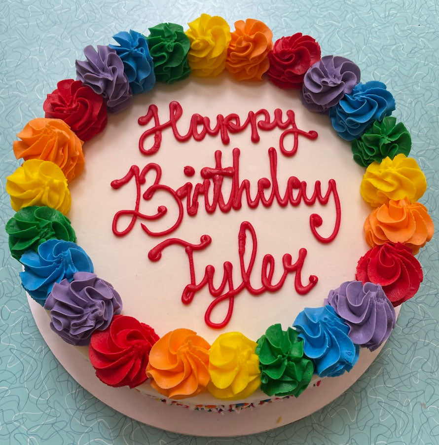 Round white birthday cake with rainbow rosettes and red icing on top with the words "Happy Birthday Tyler". 