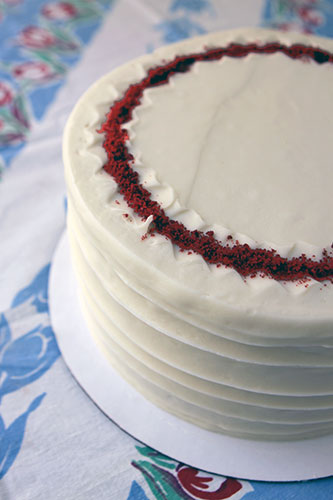 A multi-layered red velvet cake with cream cheese frosting, garnished with a ring of red velvet dust around the top perimeter, on a plate.