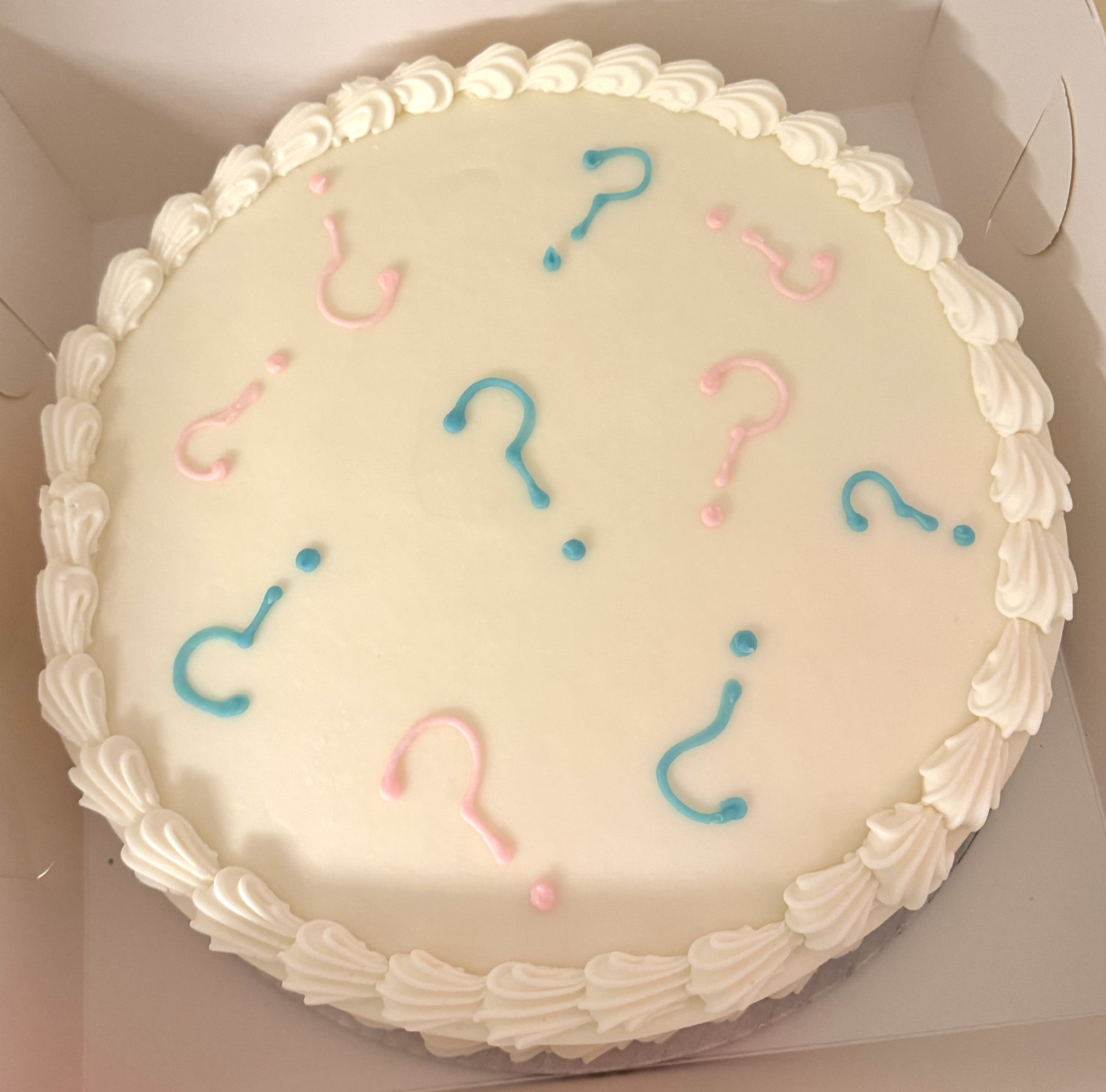 A round white cake with white buttercream and blue and pink question marks on top.