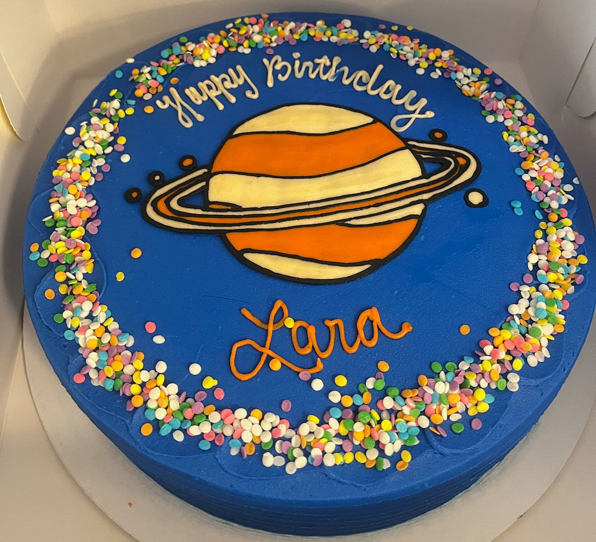 Blue birthday cake with a planet design resembling Saturn in the center, surrounded by colorful sprinkles. The cake has 'Happy Birthday' written at the top and 'Lara' written below the planet.
