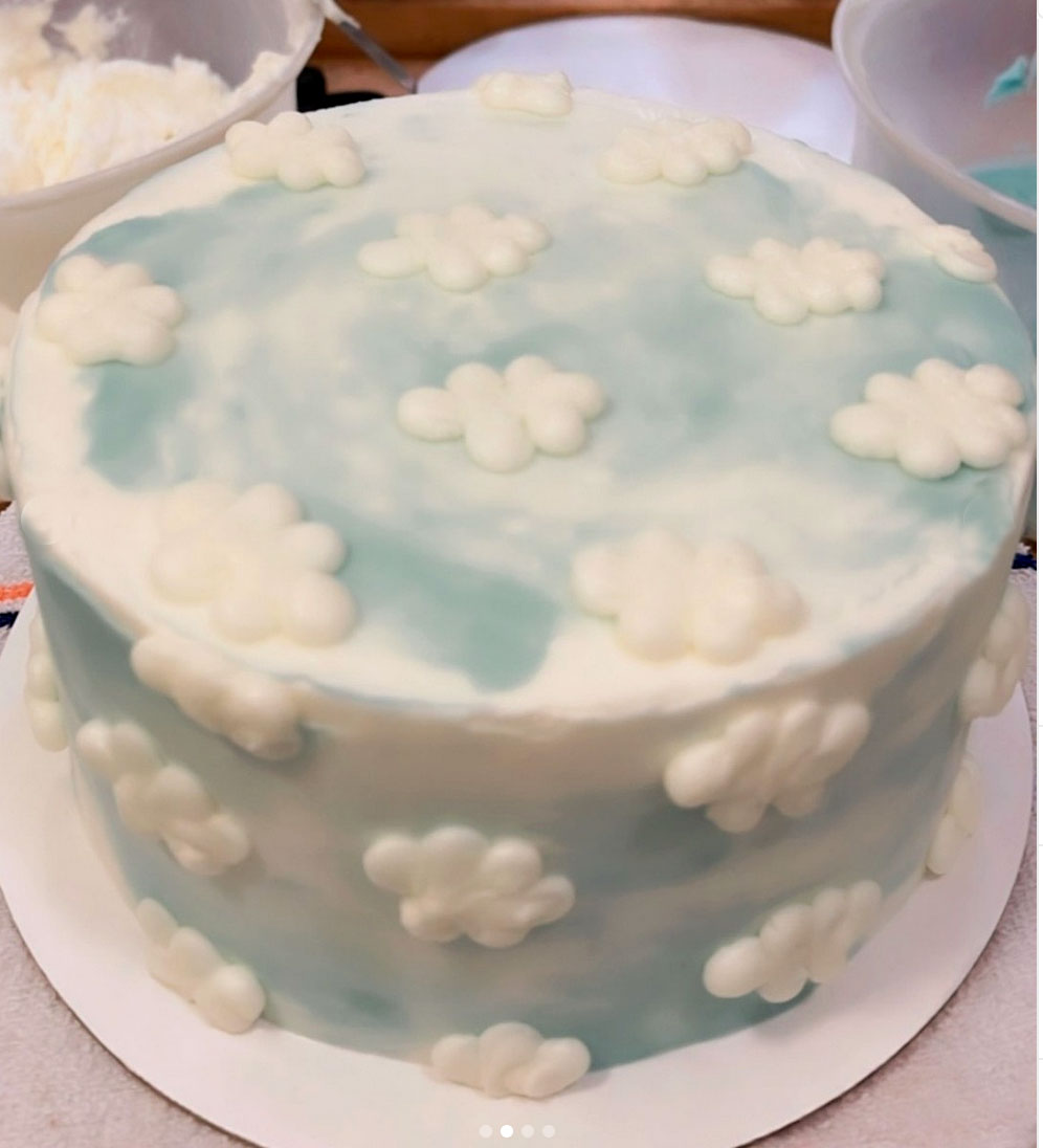 A round cake decorated with a light blue and white swirled frosting resembling a sky with white fondant clouds scattered across the top and sides.
