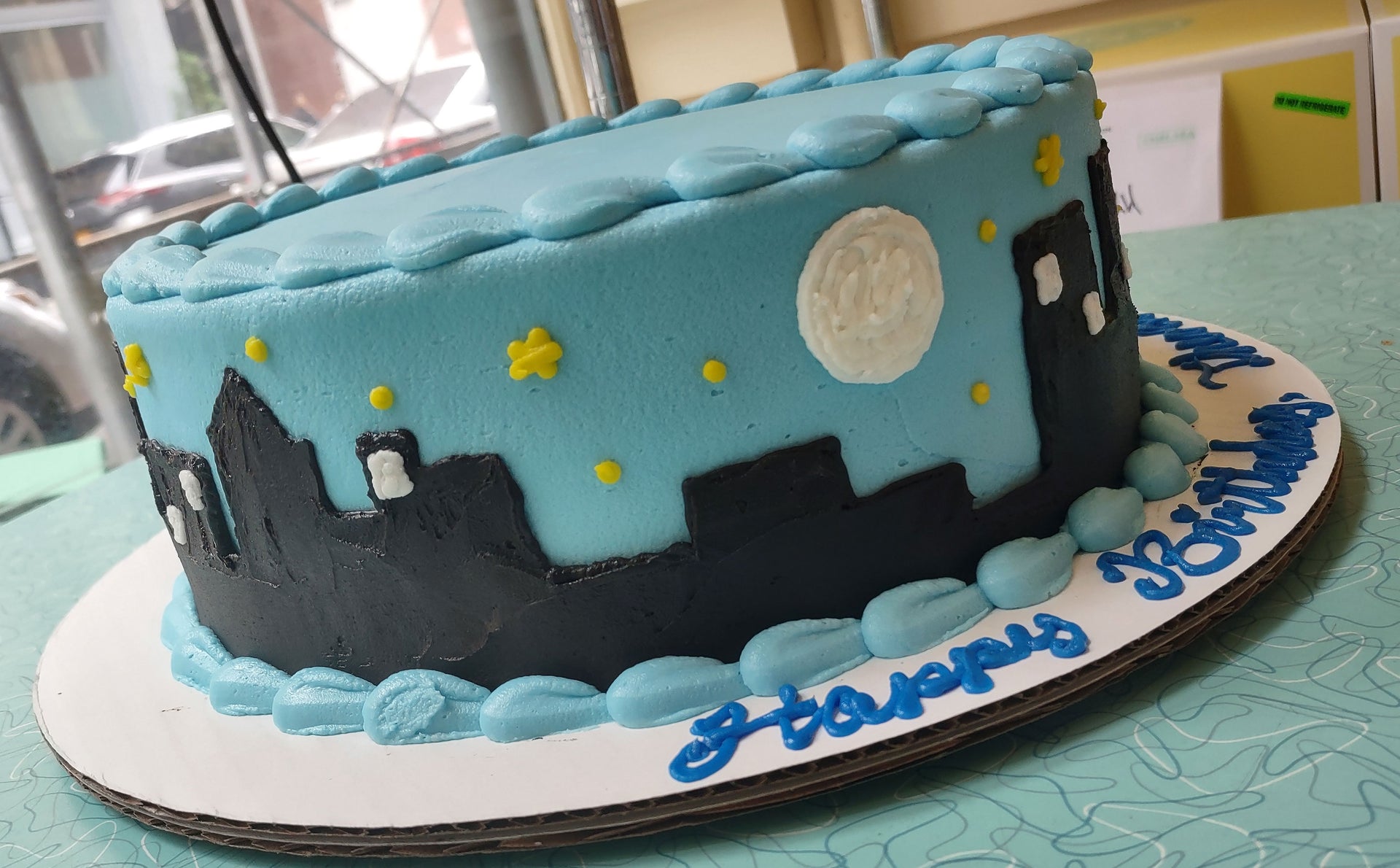 A rectangular birthday cake decorated with a blue sky, black city skyline, white full moon, and yellow stars, with 'Happy Birthday' written in blue frosting on the white cake board.