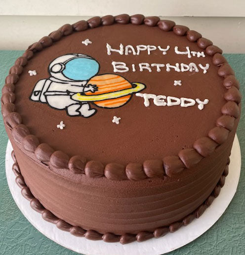 A round chocolate birthday cake with an astronaut holding a planet decorated on top and "Happy 4th Birthday Teddy" in white icing.