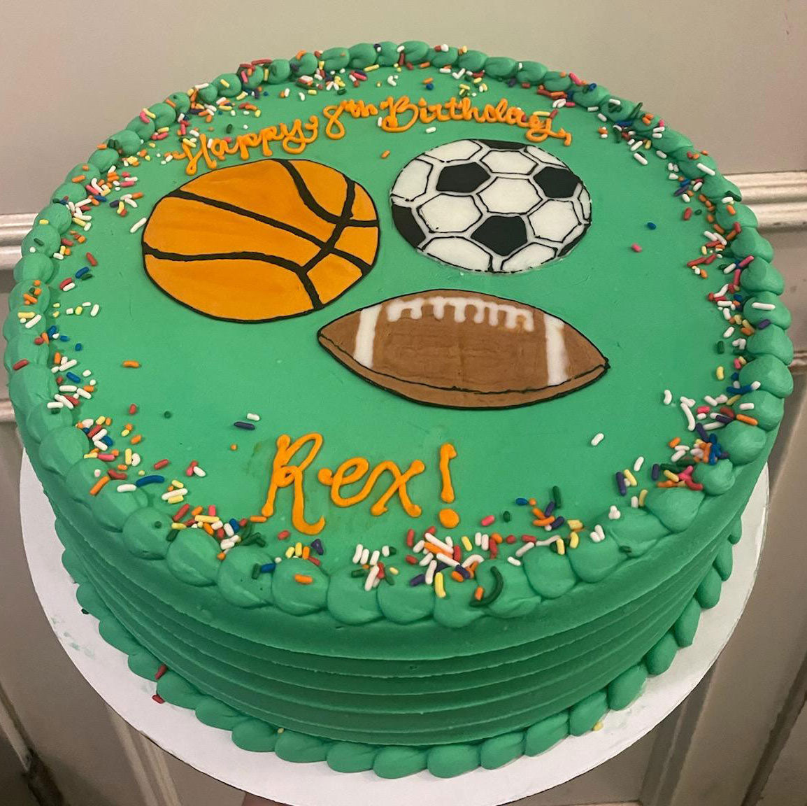 Green birthday cake for Rex with sports theme, featuring illustrations of a basketball, soccer ball, and football, and 'Happy 8th Birthday Rex!' in orange icing.